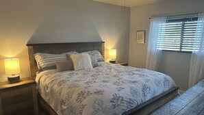 A bedroom with a large bed, two bedside tables with lamps, a wooden bench, a ceiling fan, and a window with blinds. - Waterfront Snowbird retreat with screened-in lanai and heated pool! (Cape Coral)