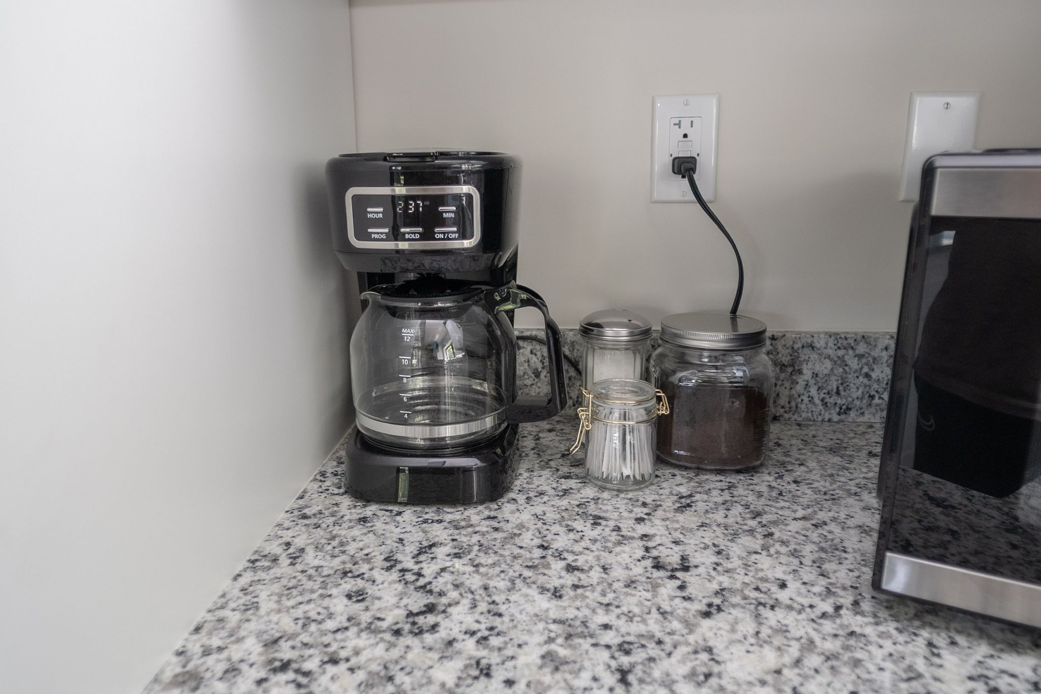 A coffee maker on a granite countertop with a glass carafe, two jars with lids, and a power outlet.