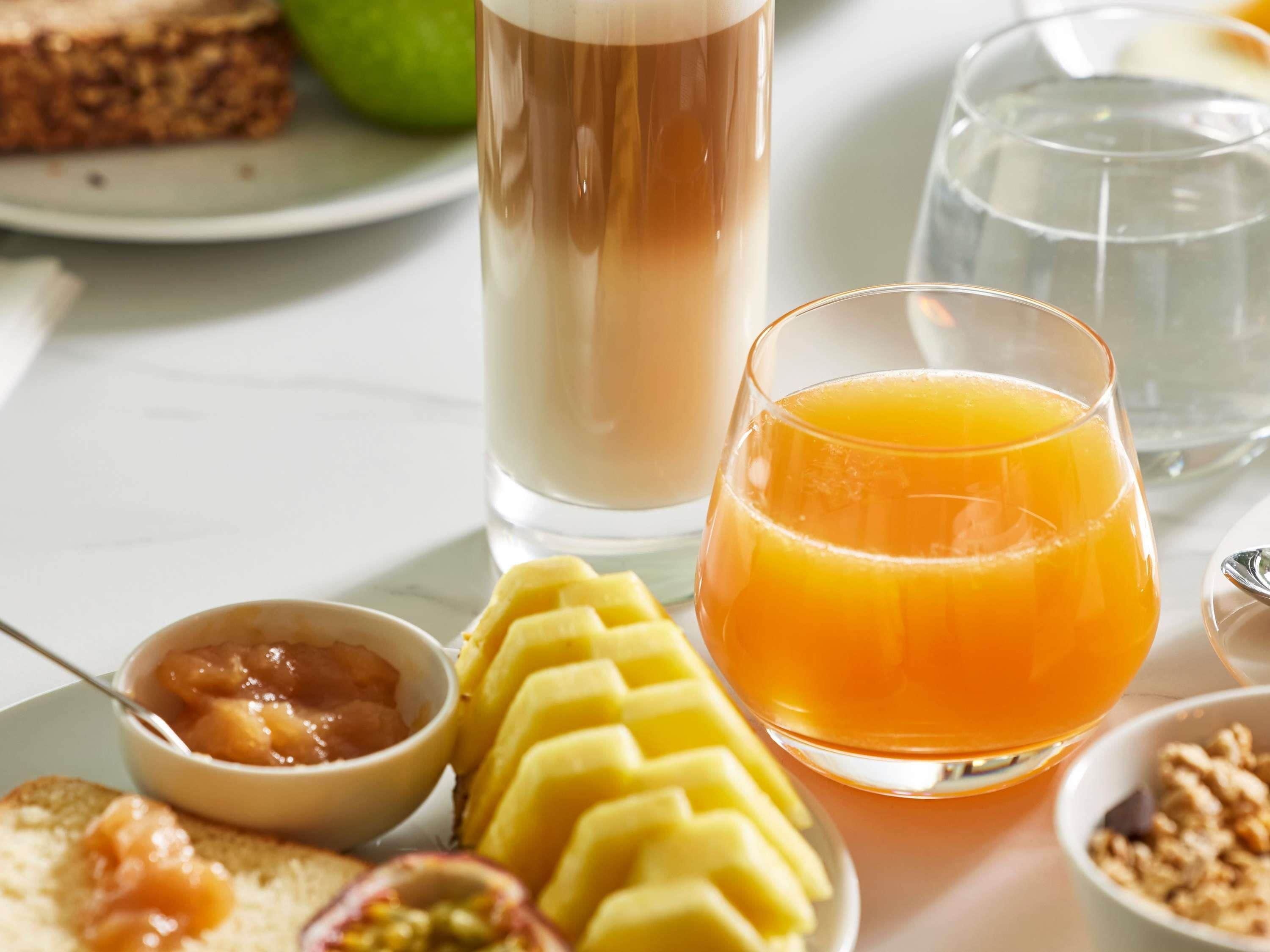 Un desayuno con un vaso de jugo de naranja, un vaso alto de una bebida marrón claro, un plato con tostadas y fruta, y un tazón de nueces.