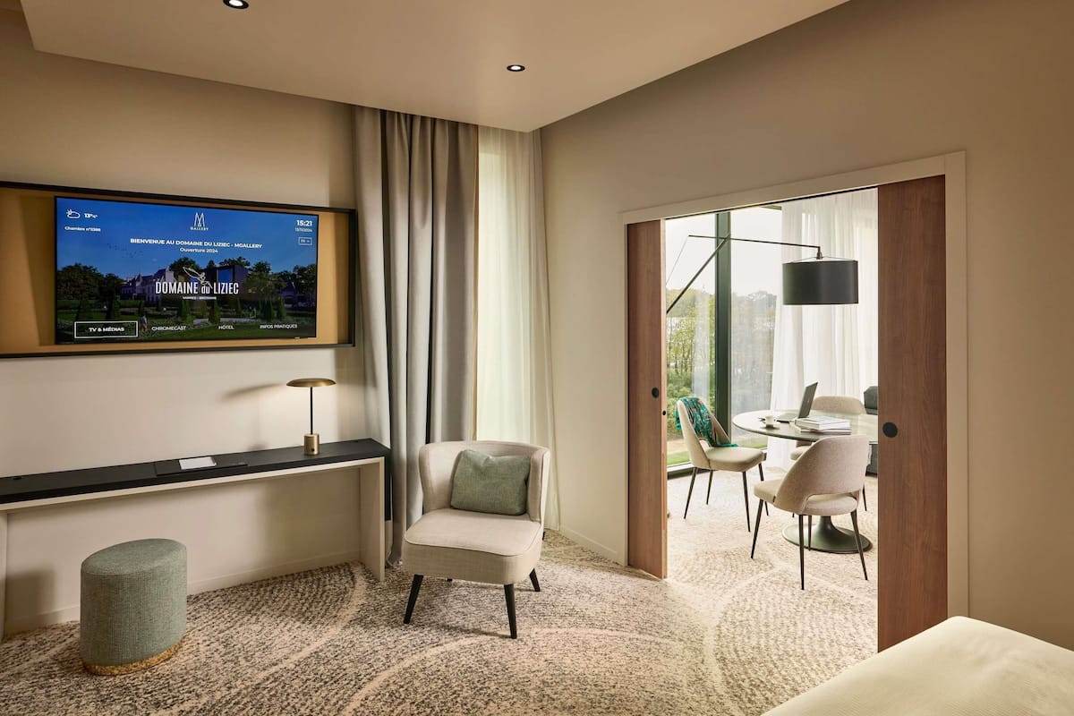 Een moderne hotelkamer met een televisie waarop een welkomstboodschap staat, een zithoek met een tafel en stoelen, en uitzicht door een glazen deur.