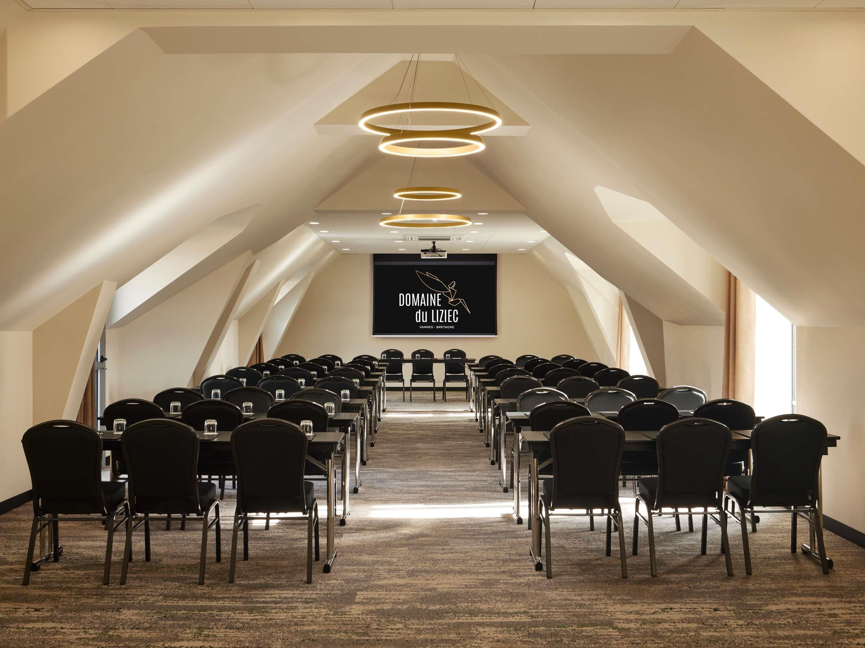 Une salle de conférence intérieure, avec des rangées de chaises, un écran de projection et un plafond orné de luminaires circulaires.