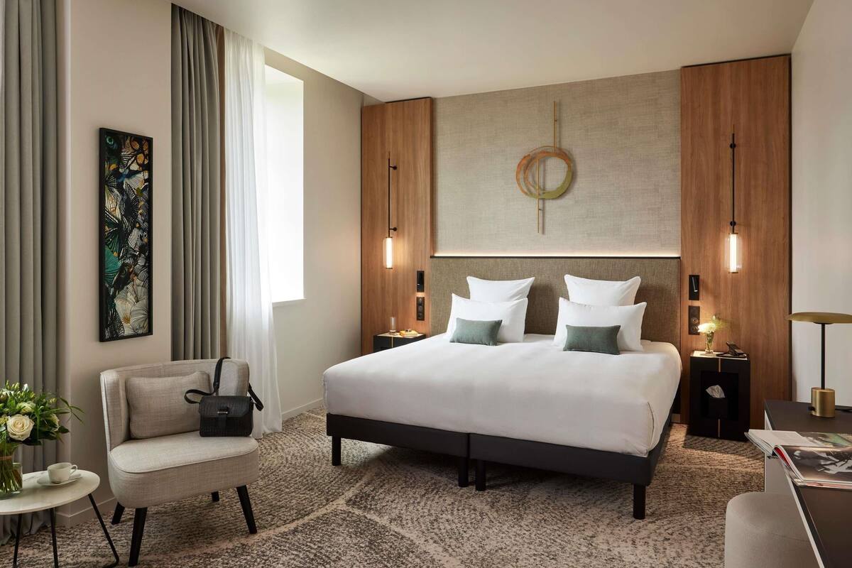 Ein modernes Hotelzimmer mit einem großen Bett, einem Sessel, einem kleinen Tisch mit Blumenvase und einem Schreibtisch mit Lampe.