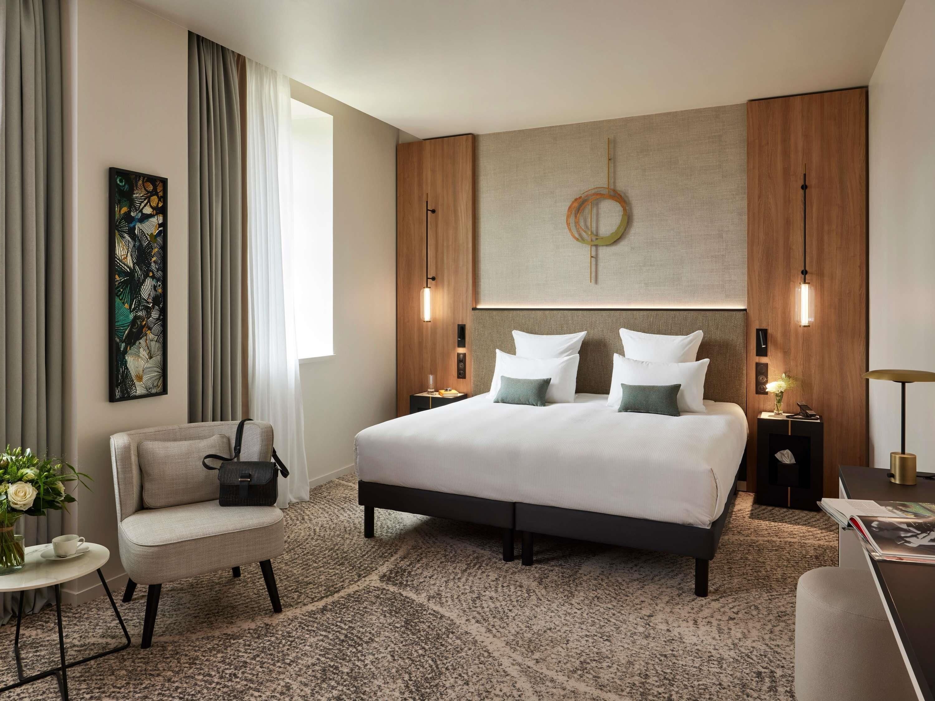 A modern hotel room with a large bed, a chair, a small table with a vase of flowers, and a desk with a lamp.