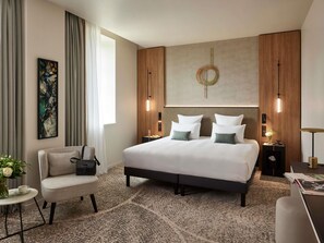 Une chambre d’hôtel moderne avec un grand lit, une chaise, une petite table ornée d’un vase de fleurs et un bureau avec une lampe.