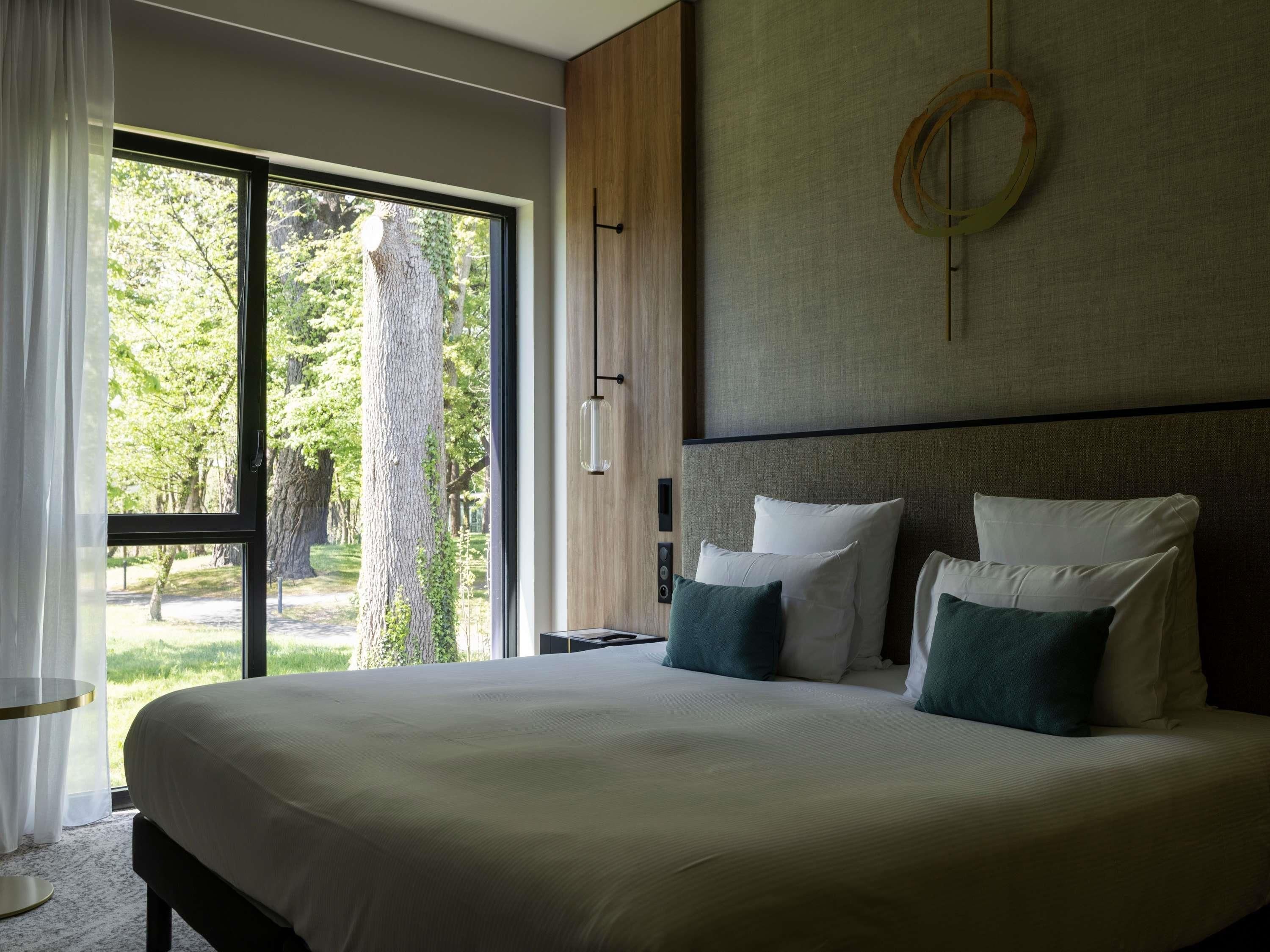 A large bed with white and green pillows, a wooden headboard, and a view of trees through a large window.
