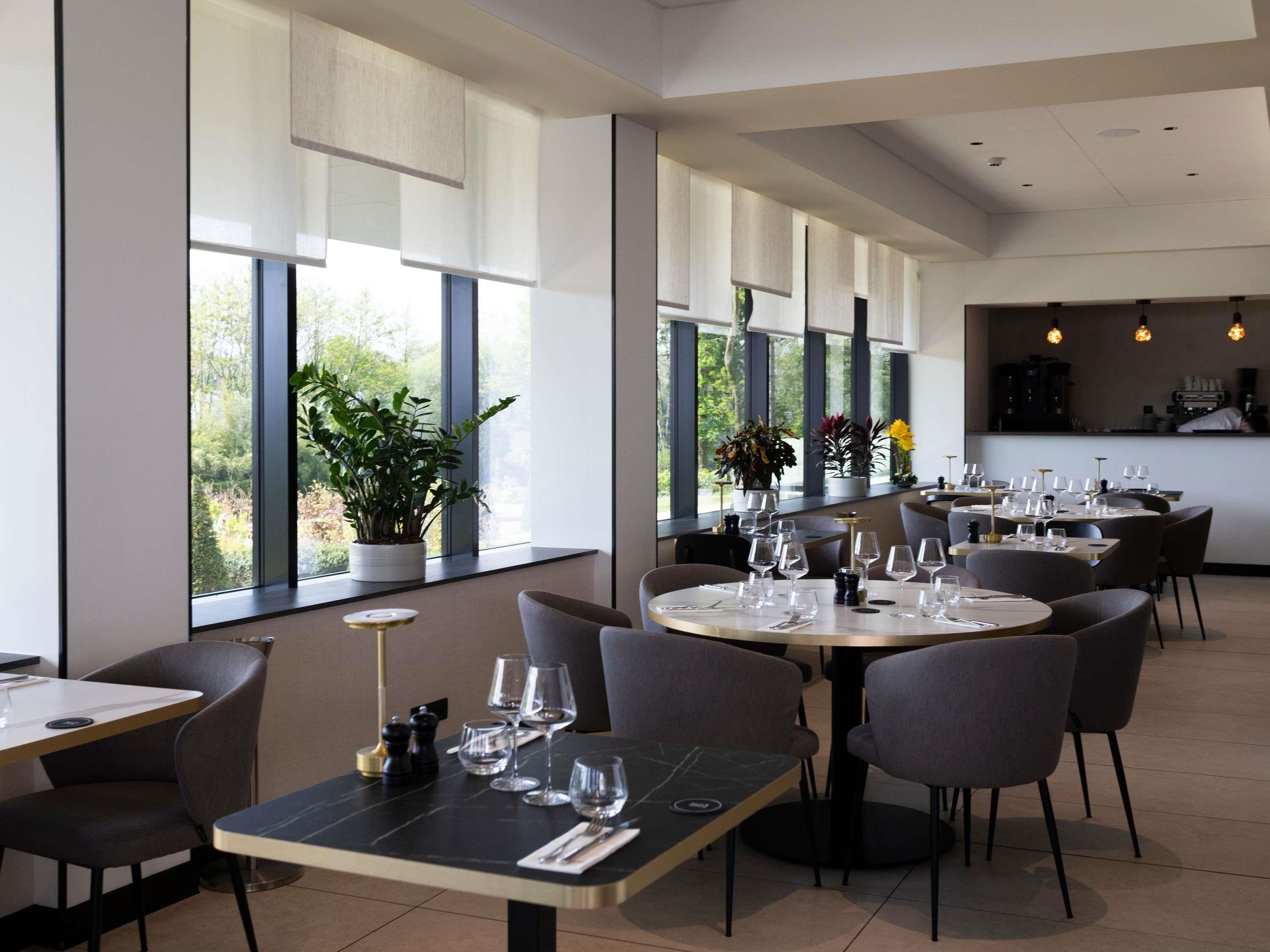 Een modern restaurant met ronde tafels, stoelen en grote ramen met uitzicht op groen.