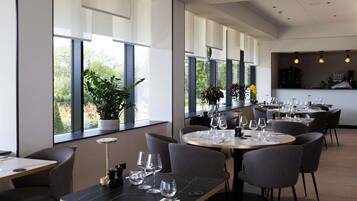 Een modern restaurant met ronde tafels, stoelen en grote ramen met uitzicht op groen.