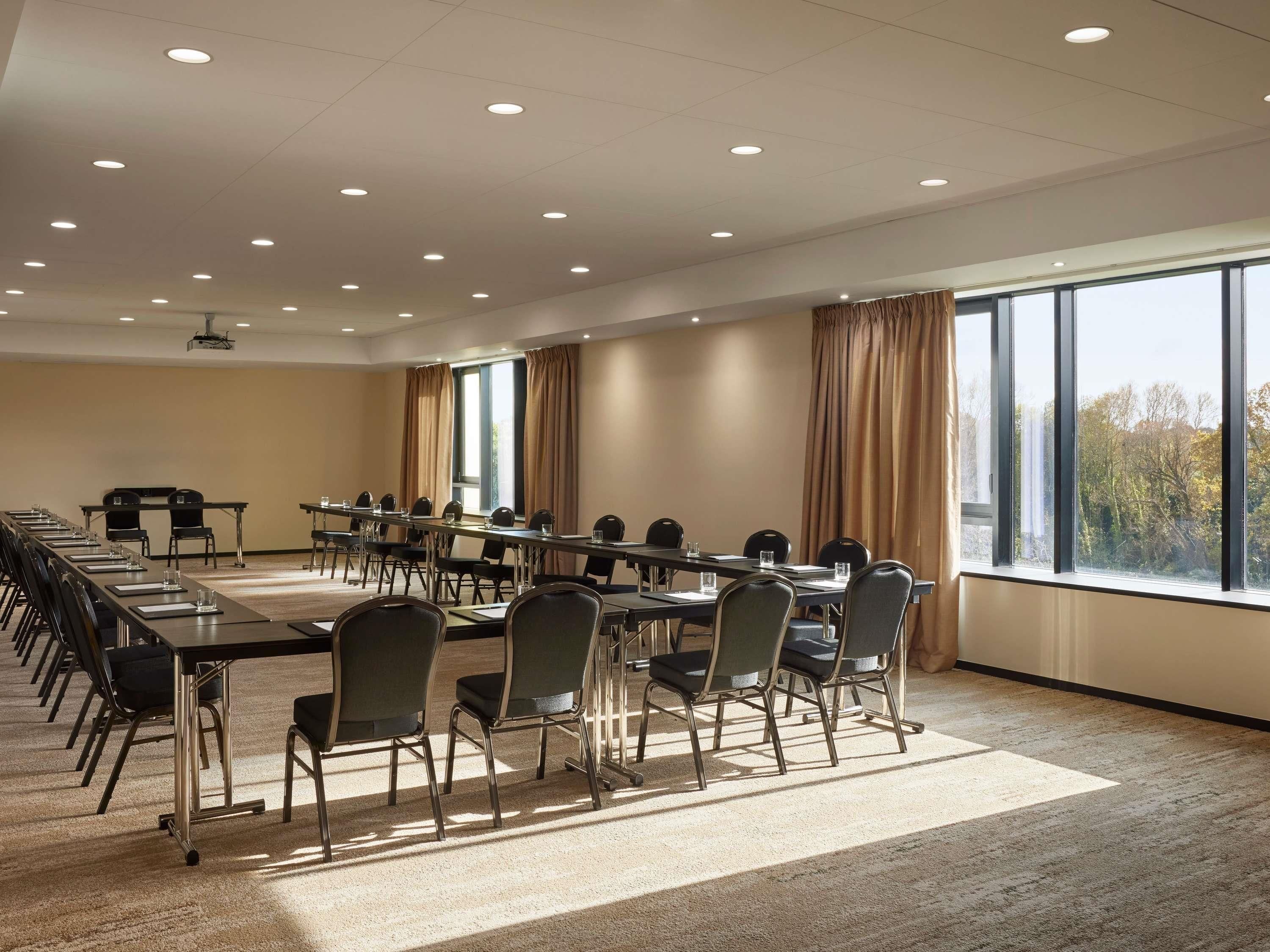 Un salón de conferencias con una larga mesa, sillas, un proyector y ventanas amplias con cortinas.
