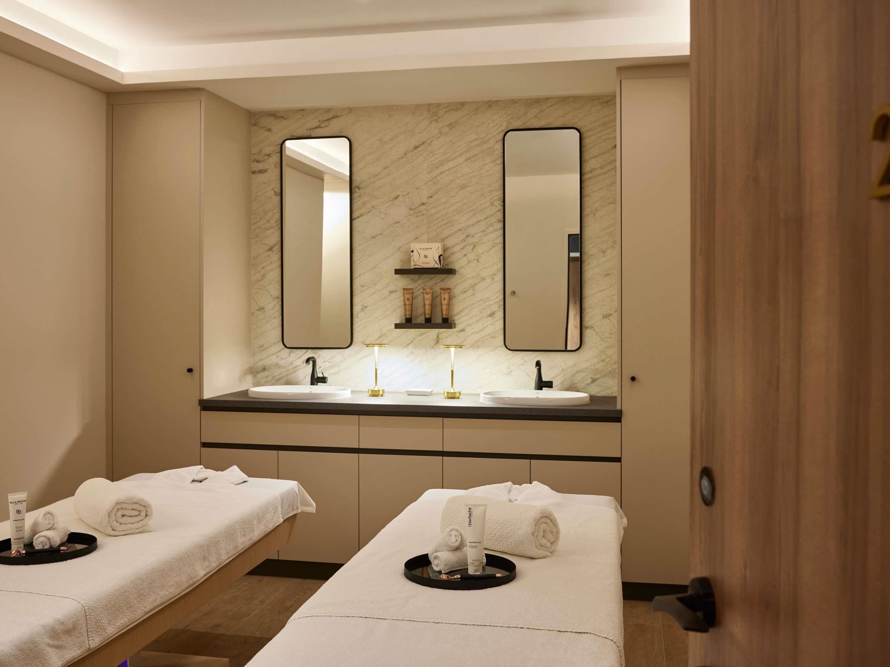 Une chambre d’hôtel avec deux lits, un grand miroir, un mur en marbre et une coiffeuse avec deux vasques.