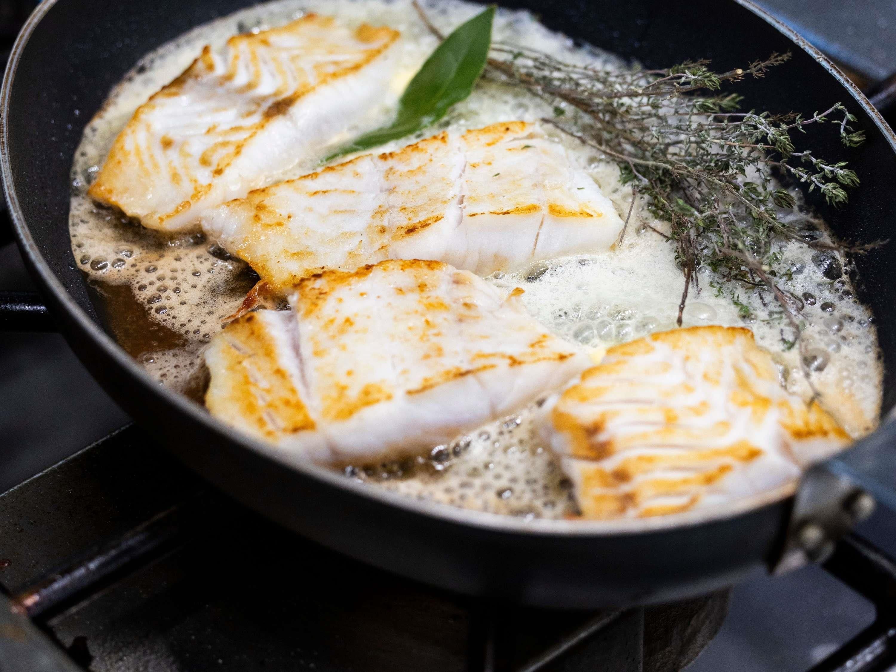 A pan with fish fillets cooking in oil, with herbs visible.