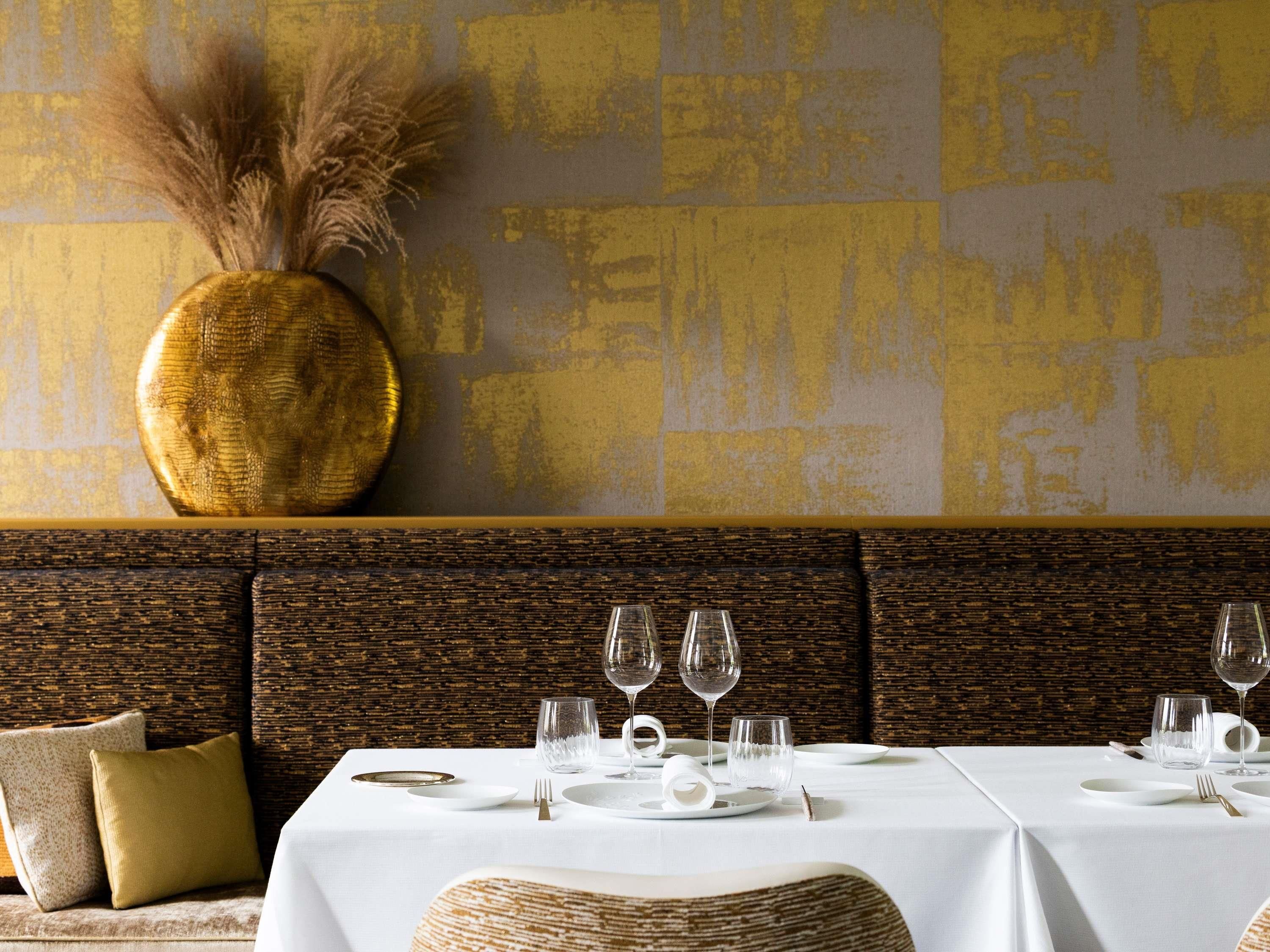Uma área de refeições com uma mesa branca posta para quatro pessoas, um vaso dourado decorativo e uma parede com textura.