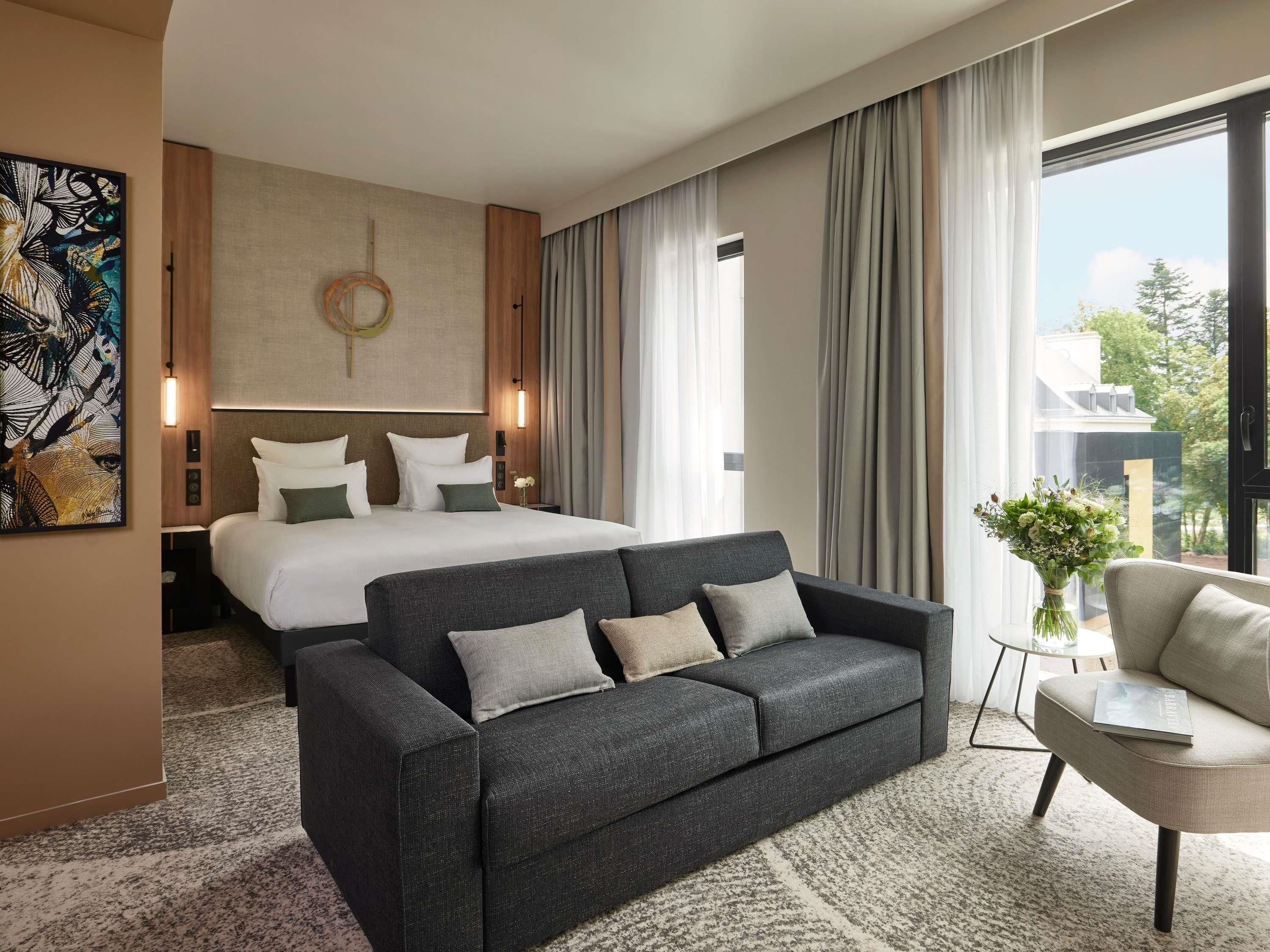 Quarto de hotel com uma cama, um sofá cinza escuro, uma mesa pequena e uma janela com cortinas.