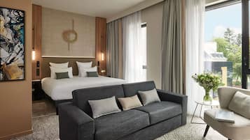 Quarto de hotel com uma cama, um sofá cinza escuro, uma mesa pequena e uma janela com cortinas.
