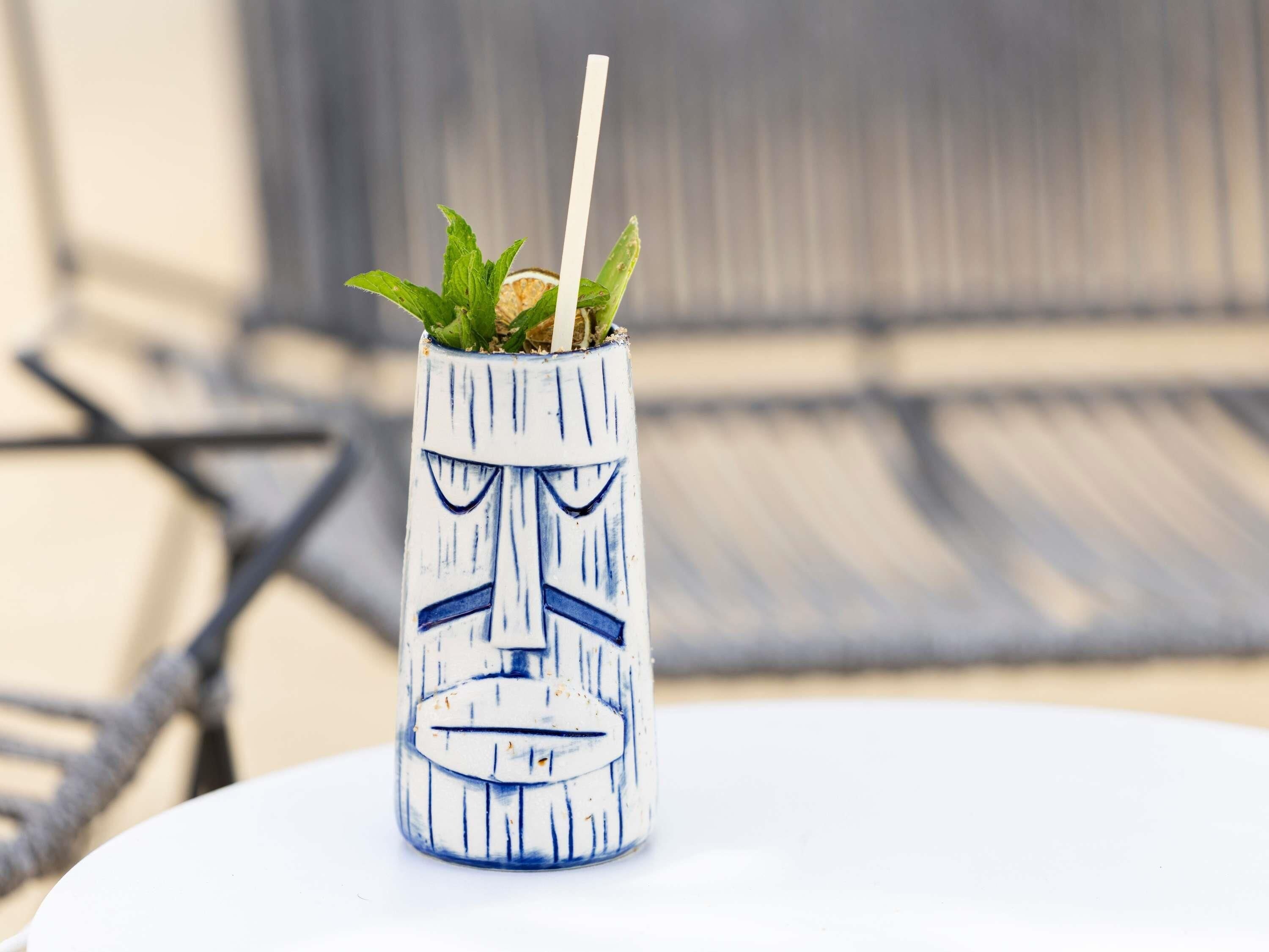 A tiki mug with a face design, a straw, and green leaves on a table.