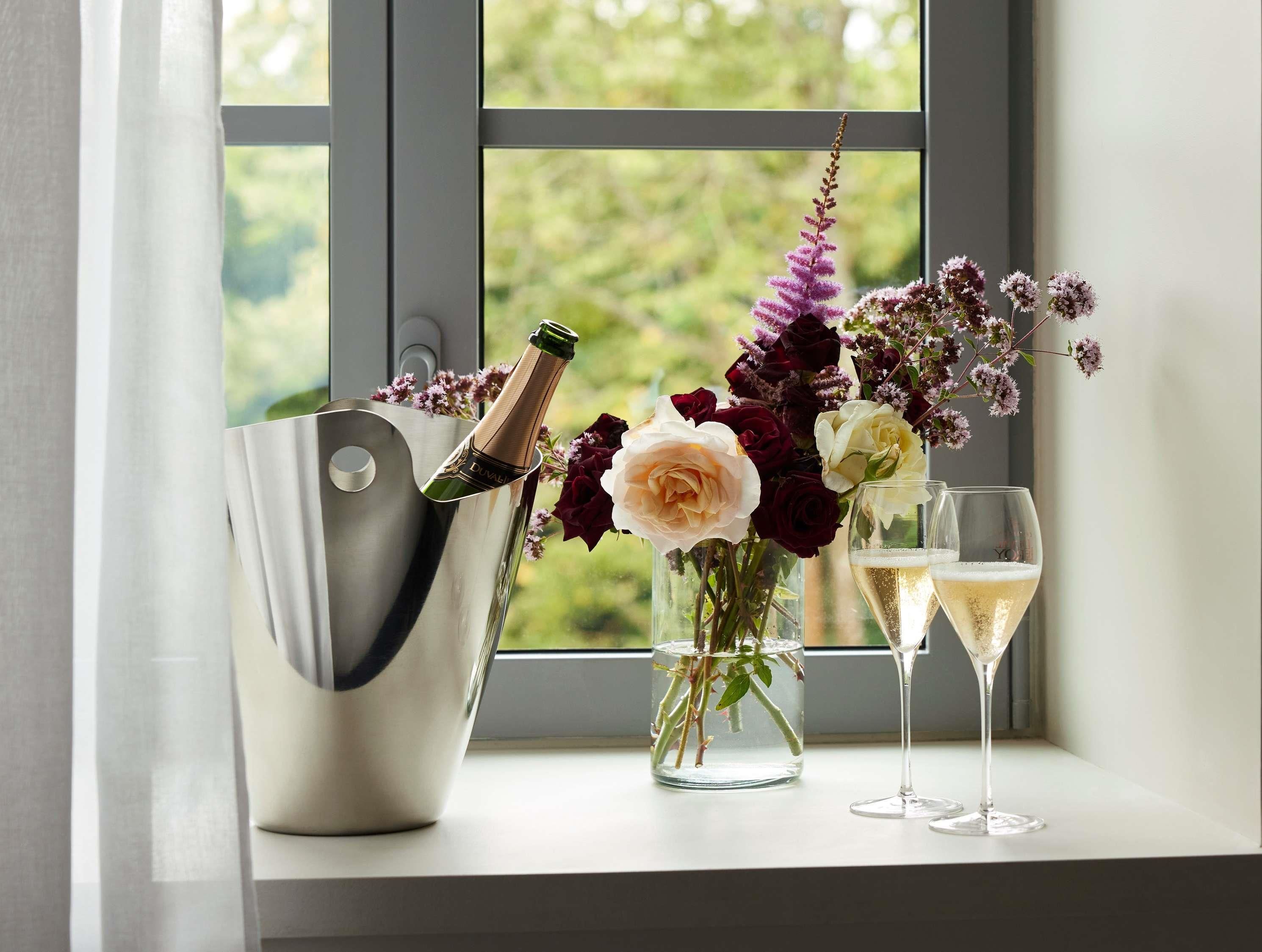 A window with a view of greenery, a champagne bottle in a metallic bucket, and two glasses of champagne on a windowsill.