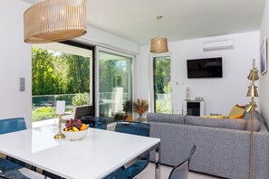 Uma sala de estar moderna com um sofá, uma mesa de jantar com uma tigela de frutas e uma TV fixada na parede.