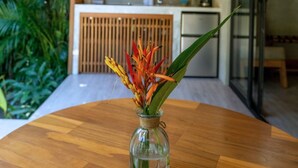 Een houten tafel met een glazen vaas gevuld met bloemen, een plafondventilator, een keuken met kasten en apparatuur, en een glazen deur die naar een andere kamer leidt.