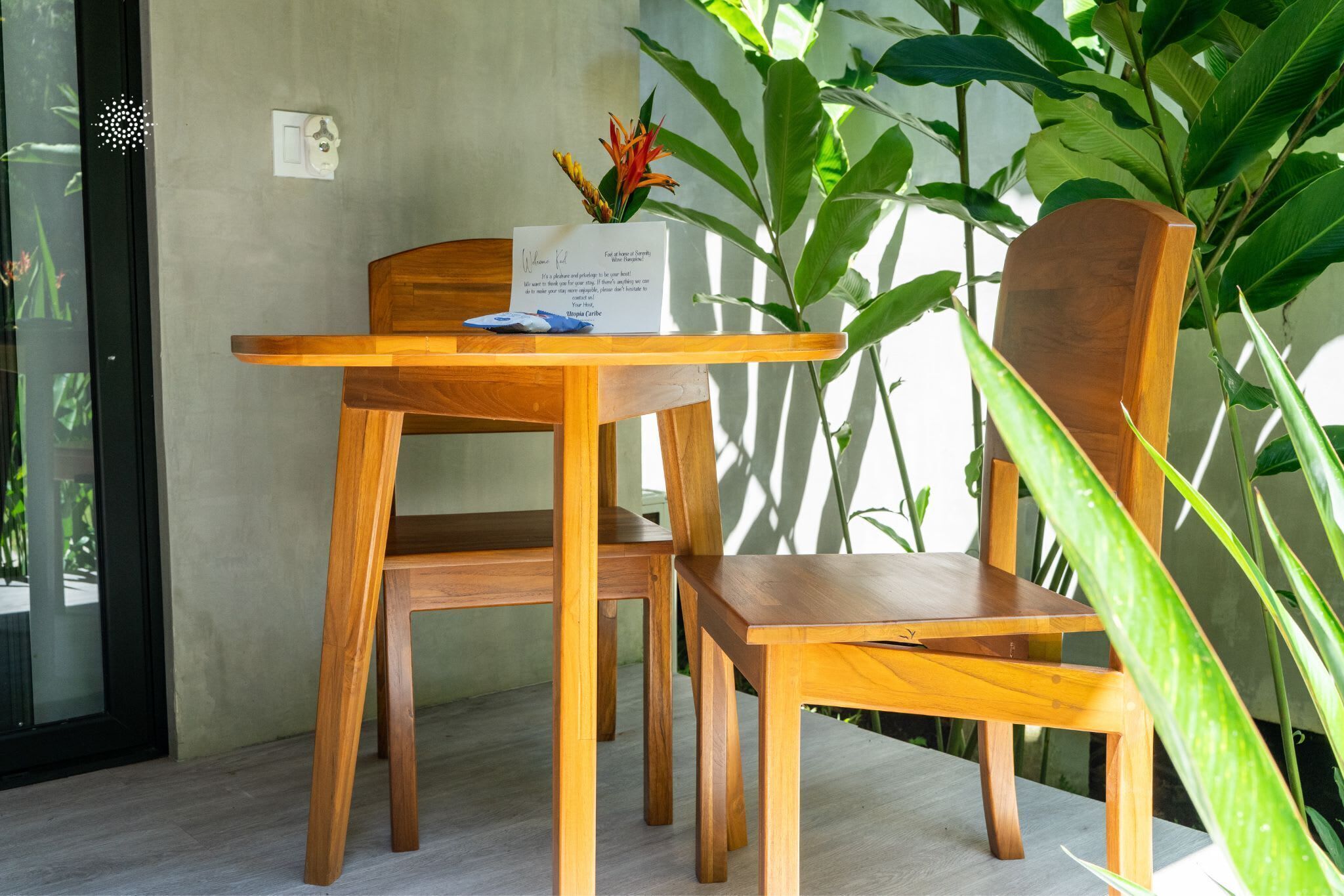 A wooden table and chairs with a welcome card, a potted plant, and a glass door.