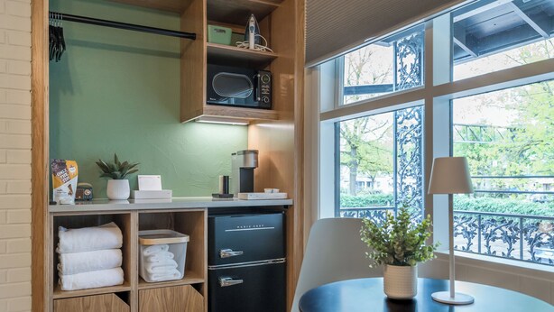 A compact kitchen with a microwave, a small refrigerator, and a window with a view of greenery. - The Local Motel & Shops (Eugene)