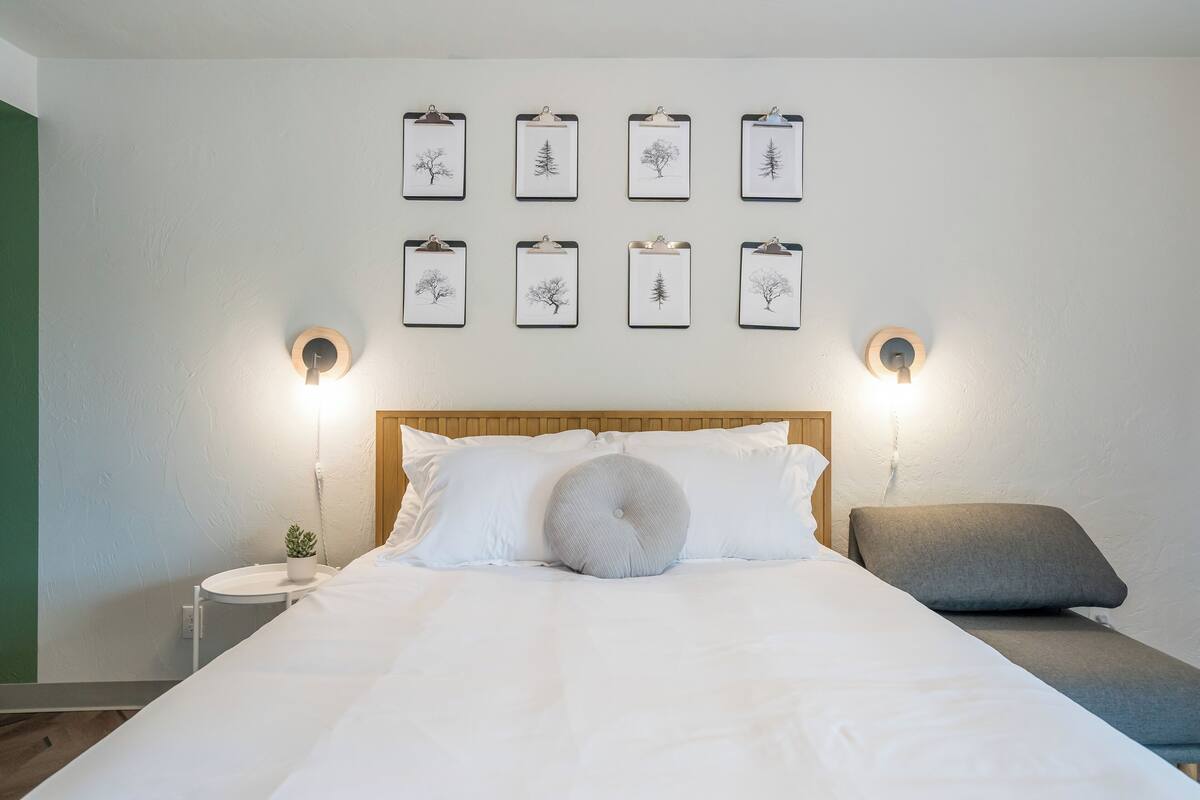 A neatly made bed with white linens, a round accent pillow, and a grey bench at the foot of the bed. There are two wall-mounted nightstands, each with a small potted plant. Above the bed, there are eight framed pictures of trees on clipboards.