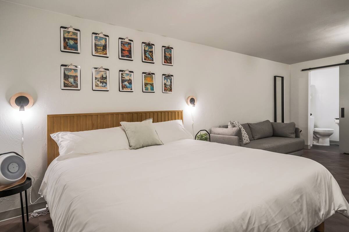A bedroom with a large bed, a wooden headboard, a grey sofa, and a wall-mounted light fixture.