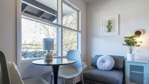 A room with a round table, two chairs, a sofa, a small cabinet, and a window with a view of trees and a fence. - The Local Motel & Shops (Eugene)
