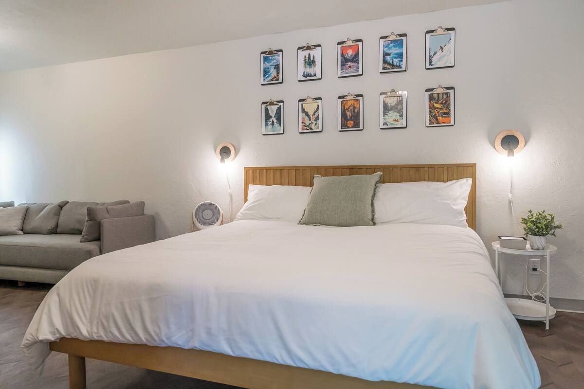 A bedroom with a large bed, a wooden headboard, a bedside table with a lamp, a wall-mounted fan, a sofa, and a series of framed pictures on the wall.
