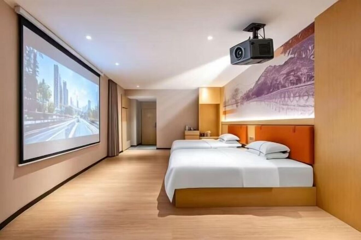 Ein Hotelzimmer mit einem großen Bett, einer Projektionsleinwand und einer Stadtlandschaftsmalerei.