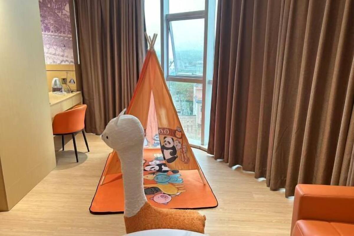 Una habitación con un ventanal grande, una zona de descanso con almohada blanca, un tapete colorido con personajes de dibujos animados y cortinas marrones.
