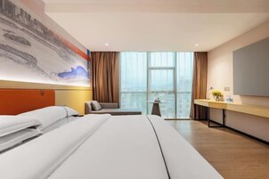 Habitación de hotel con una cama grande, un escritorio, una silla y vistas a la ciudad a través de amplias ventanas.