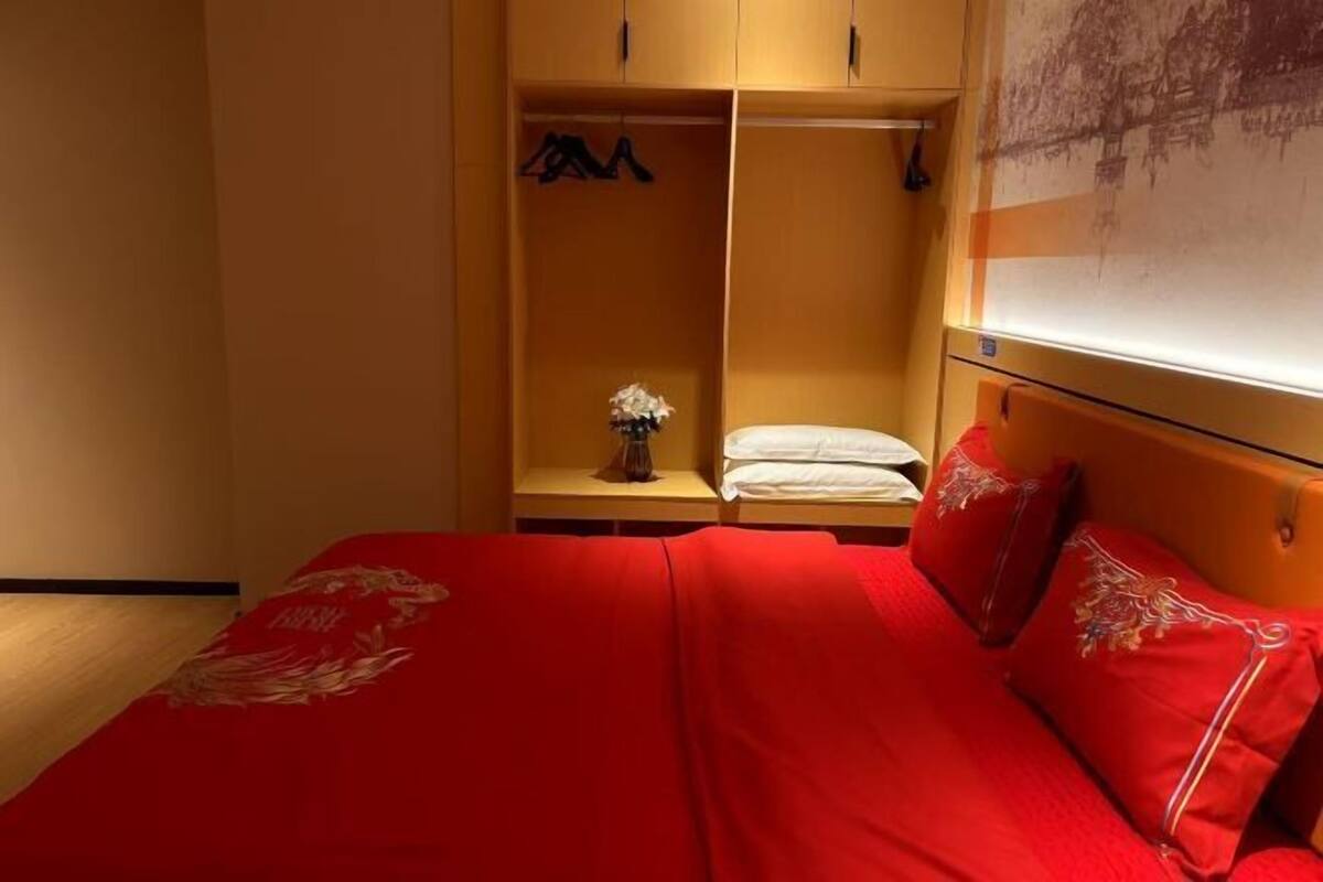 Habitación de hotel con una cama cubierta con ropa de cama roja, un armario de madera y una mesita con un jarrón de flores.