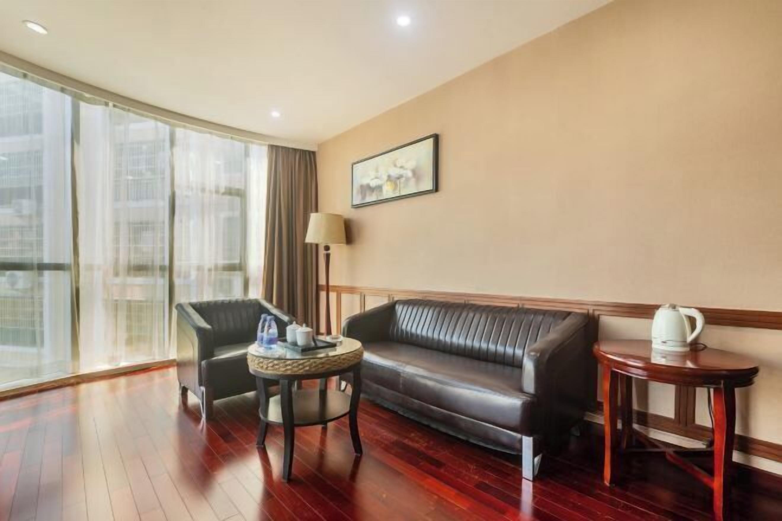 Ein Hotelzimmer mit Ledersofa, einem kleinen runden Tisch mit Lampe und Blick auf die Stadt durch große Fenster.