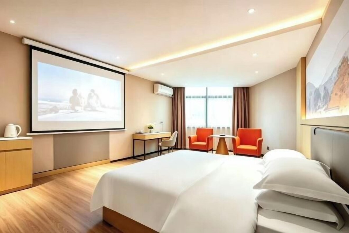 Ein Hotelzimmer mit einem großen Bett, einem Schreibtisch mit Stuhl, einem kleinen Tisch und einer Projektionsleinwand.