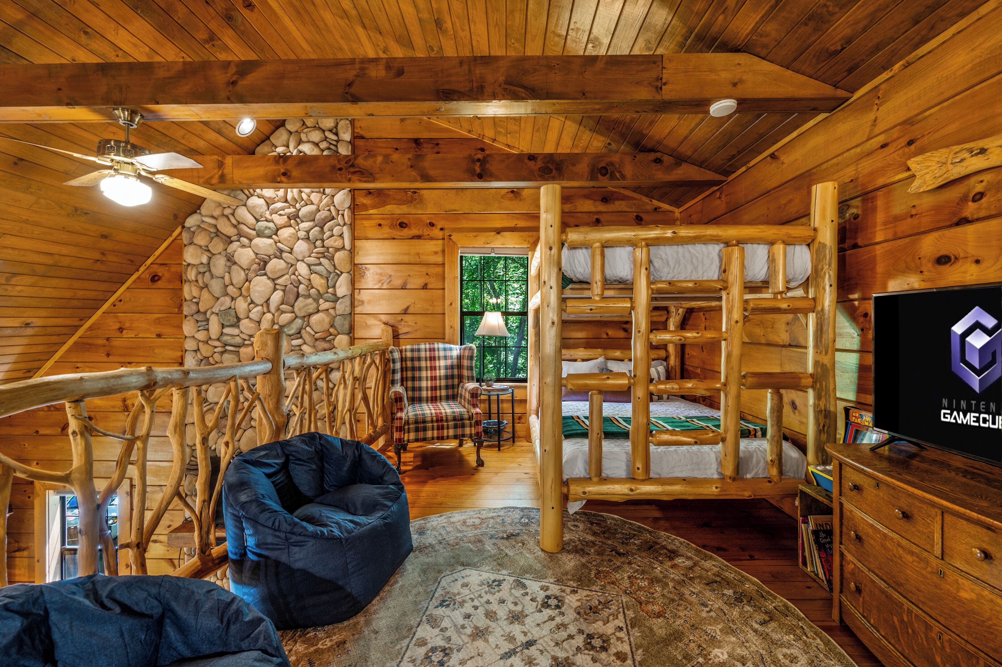 A log cabin interior with a bunk bed, a ceiling fan, a stone wall, a plaid chair, and a television.