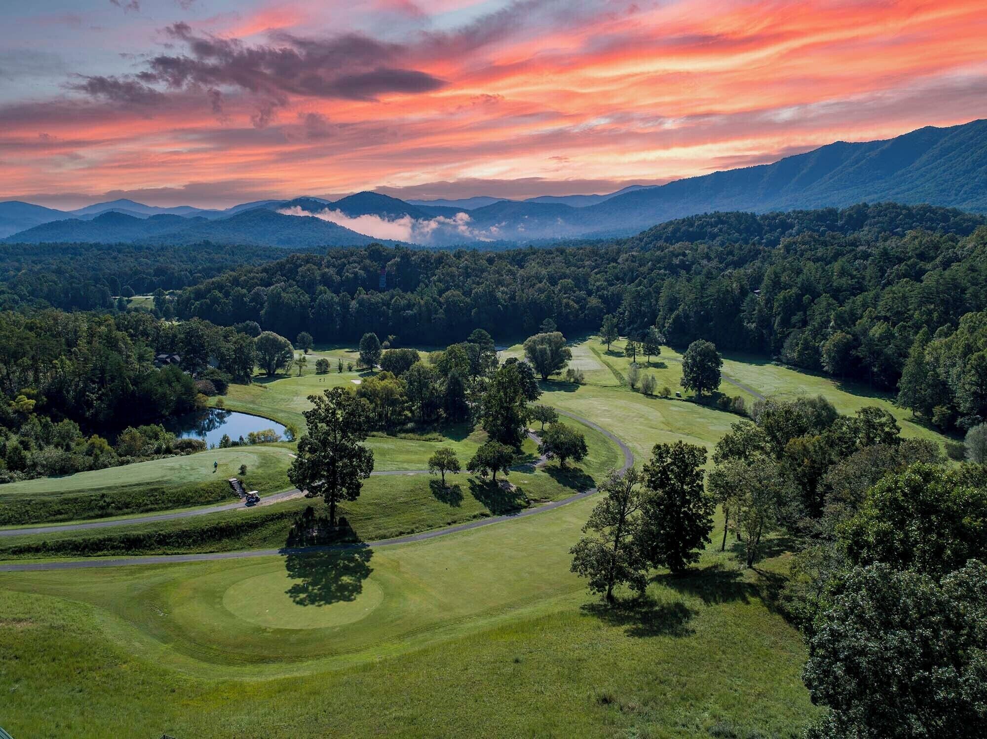 A golf course with a pond, surrounded by dense forests and mountains.