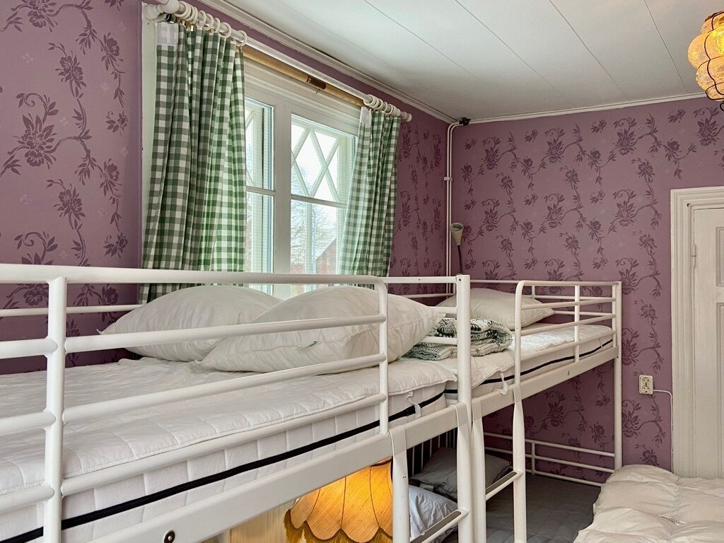A bunk bed room with floral wallpaper, a window with checkered curtains, and a lamp on the floor.