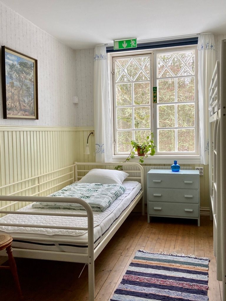 A single-bed room with a window, a bedside table, a chair, and a radiator.