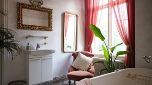 A bathroom with a white sink, a mirror with a gold frame, a red chair with a pillow, and a large window with red curtains. - Herrgård de luxe (Tävelsås)