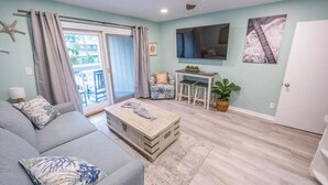 A living room with a sofa, a coffee table, a TV, a dining area with stools, and a door. - Coligny Beachfront Villa w/Pool at The Breakers (Hilton Head Island)