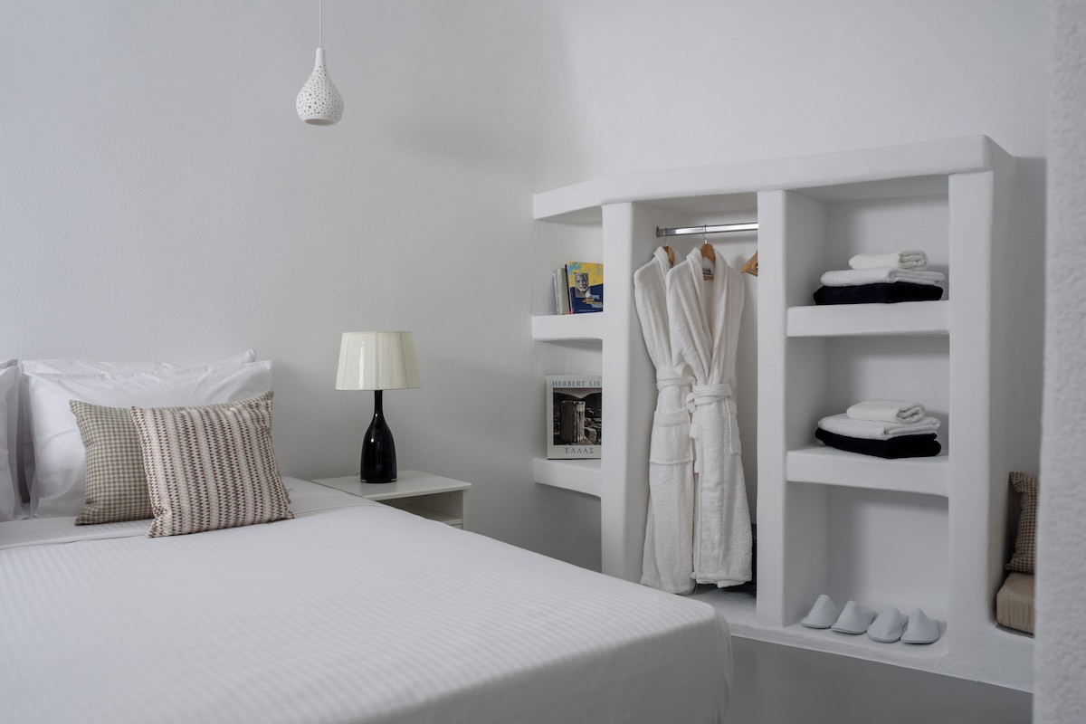 A neatly arranged hotel room with a bed, a bedside table with a lamp, a robe hanging on a rack, and a shoe shelf.