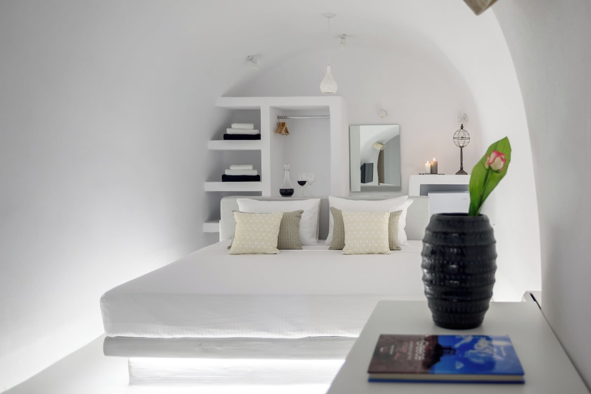 A modern bedroom with a white bed, a bedside table with a book, a black vase with a plant, and a white wardrobe with shelves.