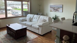 Een woonkamer met een witte bank, een houten salontafel, een eettafel en een keukenhoek met een spoelbak en planten.