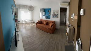 Una sala moderna con un sofá marrón, un comedor con una mesa y sillas blancas, y una cocina con armarios azules y una lámpara colgante.