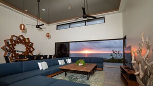 A modern living room with a large blue sectional sofa, a wooden coffee table, and a view of the sunset over the ocean. - Casa Ola Vista by Pura Villas (Dominical)