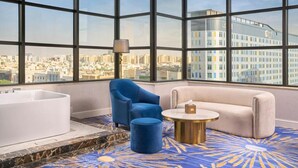 A modern hotel room with a large window, a white bathtub, a blue armchair, a beige sofa, and a round coffee table. - Novo Hotel (Riyadh)