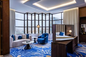 A hotel room with a large window, a sofa, a chair, a bed, a desk, and a mirror.