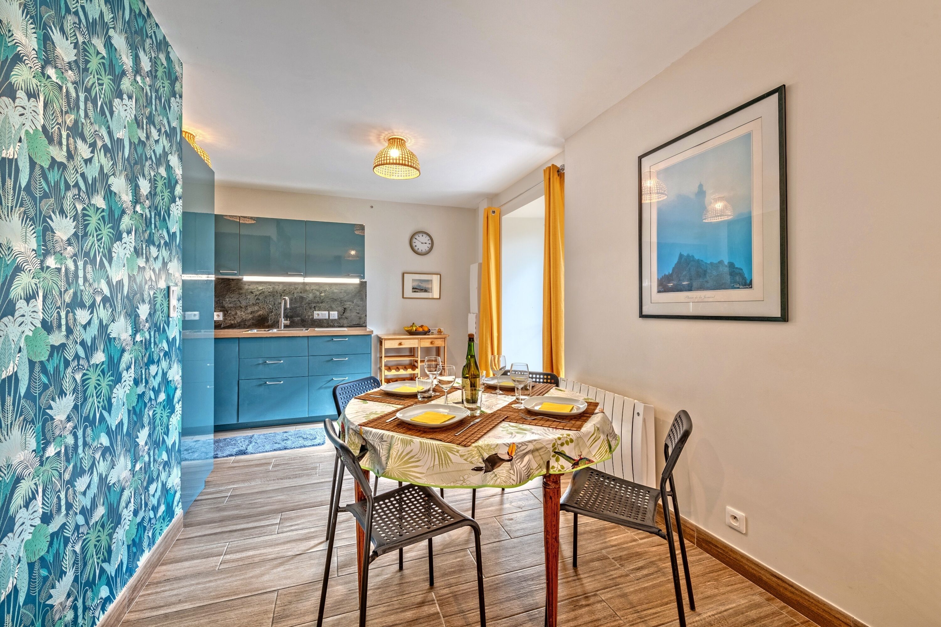 A dining area with a round table, chairs, and a bottle of wine. There is a kitchen in the background with blue cabinets and a clock on the wall.
