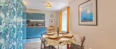 A dining area with a round table, chairs, and a bottle of wine. There is a kitchen in the background with blue cabinets and a clock on the wall.