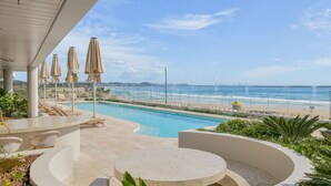 Ein Poolbereich mit Blick auf Strand und Meer, Liegestühle und Sonnenschirme.
