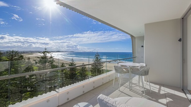 Ein Balkon mit Blick auf Strand und Meer, mit Glastrennwand und Tisch mit Stühlen.
